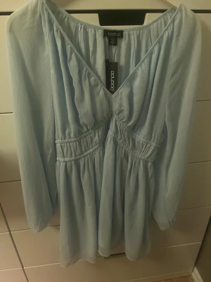 Ljusblå kortklänning från Boohoo - Supersöt ljusblå klänning från Boohoo med v-ringad hals och långa ärmar. Klänningen har snygga rynkade detaljer vid midjan och ett flowigt tyg som ger en luftig känsla. Perfekt för dig som gillar en feminin och trendig look.
