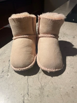 Ljus rosa UGG boots för barn - Säljer ett par supermysiga beige UGG boots för barn med mjukt pälsfoder och synliga sömmar. Skorna har rund tå och är tillverkade i mocka med UGG-logga på sidan. Perfekta för kalla dagar och ger en riktigt cozy vibe.