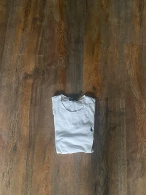 Vit t-shirt från Polo Ralph Lauren - Vit t-shirt från Polo Ralph Lauren med klassisk rund hals och korta ärmar. På bröstet finns den ikoniska broderade polospelaren i mörkblått. T-shirten är gjord i mjuk bomull och har en clean, stilren look som funkar till allt.