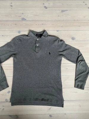 Grå långärmad pikétröja Polo Ralph Lauren - Säljer en stilren grå långärmad pikétröja från Polo Ralph Lauren. Tröjan har klassisk krage, två knappar framtill och den ikoniska broderade loggan på bröstet. Perfekt för dig som gillar enkel och tidlös design.