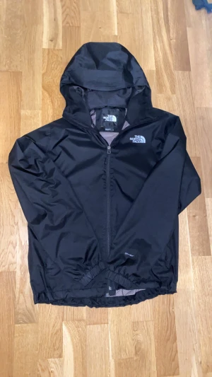 Svart vindjacka från The North Face - Svart vindjacka från The North Face med huva och hel dragkedja framtill. Jackan har meshfoder på insidan och elastiska muddar vid midja och ärmslut. Perfekt för blåsiga dagar och outdoor-aktiviteter. Snygg logga på bröstet och lätt material.