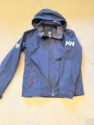Blå vindjacka från Helly Hansen - Snygg marinblå vindjacka från Helly Hansen med huva och röda detaljer på dragkedjorna. Jackan har lång ärm, HH-logga på bröstet och är tillverkad i slitstark polyester. Perfekt för blåsiga dagar och outdoor-aktiviteter.