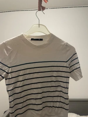 Vit randig t-shirt från 157 - Säljer en fräsch vit t-shirt från 157 i storlek S med tunna svarta  horisontella ränder. T-shirten har rund hals och korta ärmar, perfekt för en chill stil. Materialet känns mjukt och lätt, troligen bomull. Enkel och snygg design som funkar till jeans eller shorts.