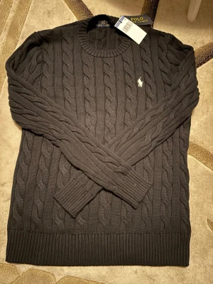 Mörkgrå stickad tröja Polo Ralph Lauren - Snygg mörkgrå stickad tröja från Polo Ralph Lauren i storlek M. Tröjan har ett klassiskt kabelstickat mönster, rund hals och långärmade ribbade muddar. Ikoniska Polo-loggan broderad på bröstet. Perfekt för dig som gillar stilren och tidlös design.