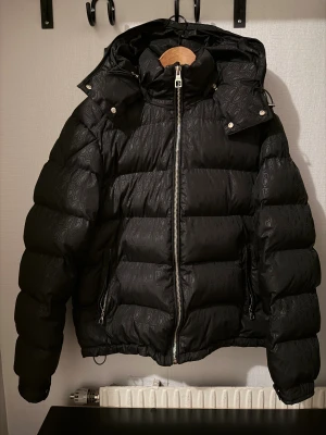 Svart pufferjacka från banditeurope - Säljer en svart pufferjacka från banditeurope med huva och coolt mönster med B-loggor över hela jackan. Jackan har dragkedja framtill, två sidofickor med dragkedja och detaljer med knappar vid huvan. Perfekt för kalla dagar och riktigt snygg streetstil.