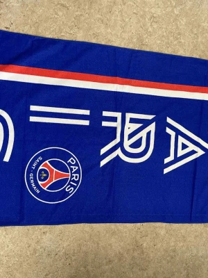 Blå PSG väst med tryck - Säljer en blå väst från Paris Saint-Germain med vitt och rött tryck längs bröstet och PSG-loggan på framsidan. Västen har en sportig look och är perfekt för dig som gillar fotboll och vill visa vilket lag du hejar på.