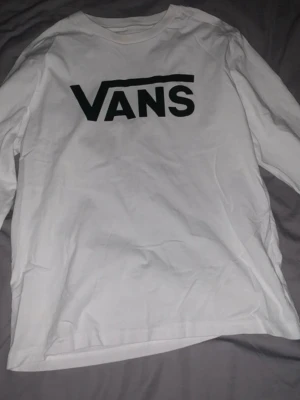 Vit långärmad tröja från Vans - Säljer en vit tröja från Vans med stor svart logga på bröstet. Klassisk rund hals och rak passform. Perfekt för dig som gillar streetwear och vill ha en enkel men snygg look.
