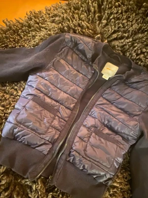 Mörkblå kofta från Gina  - Mörkblå kofta från Gina Tricot i storlek 158/164. Jackan har glansiga paneler framtill, två fickor med lock och ribbade muddar vid ärmslut och nederkant. Dragkedja hela vägen och mjuka stickade ärmar för extra komfort.