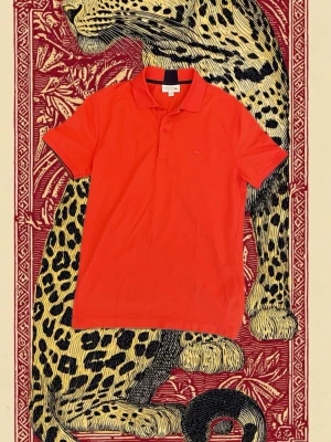 Röd pikétröja från Lacoste - Säljer en snygg röd pikétröja från Lacoste i regular fit. Tröjan har klassisk krage, korta ärmar och två knappar framtill. Materialet är bomull och den har den ikoniska Lacoste-krokodilen ton-i-ton på bröstet. Perfekt för dig som gillar stilrena och sportiga plagg.