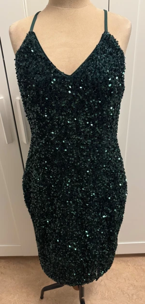 Mörkgrön glittrig klänning från SHEIN - Supersnygg mörkgrön kortklänning från SHEIN med glittriga paljetter över hela klänningen. Den har smala axelband, v-ringad fram och dragkedja bak. Perfekt för dig som vill sticka ut med en festlig look. Materialet är syntet och klänningen har en tight passform.