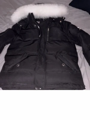 Svart Moose Knuckles dunjacka med päls - Svart dunjacka från Moose Knuckles med vit fluffig päls på huvan och ikonisk logga på ärmen. Jackan har flera fickor med dragkedja och knappar, samt quiltad design som ger extra värme. Perfekt för kalla dagar och riktigt snygg streetstil.