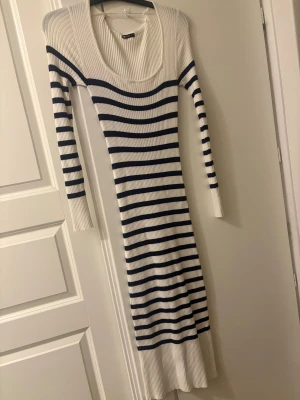 Randig långklänning från Gina Tricot - Säljer en ribbad långklänning från Gina Tricot i storlek S. Klänningen är vit med marinblå horisontella ränder, har lång ärm och en djup rundad halsringning. Materialet är stickat och ger en slim passform som sitter snyggt på kroppen.