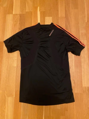Svart Adidas t-shirt med orange ränder - Säljer en svart Adidas t-shirt med klassiska orange ränder längs axlarna och ärmarna. Tröjan har Adidas-loggan på bröstet och coola orange detaljer på framsidan. Perfekt för träning eller sport, tillverkad i lätt syntetmaterial som andas.
