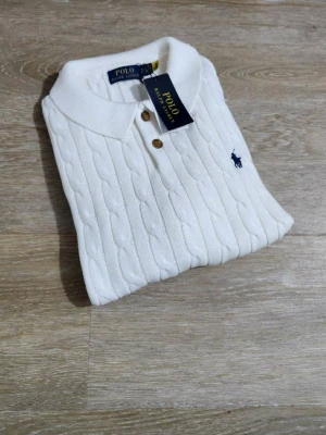 Vit kabelstickad pikétröja Polo Ralph Lauren - Vit kabelstickad pikétröja från Polo Ralph Lauren med klassisk krage, korta ärmar och bruna knappar framtill. Tröjan har broderad marinblå logga på bröstet och är gjord i mjuk bomull. Perfekt för en clean och stilren look.