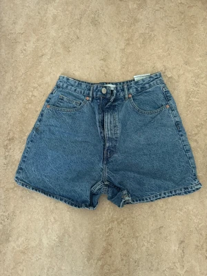 Högmidjade jeansshorts från Zara - Säljer ett par klassiska blå jeansshorts från Zara med hög midja och fem fickor. Shortsen har en loose passform, knapp och dragkedja framtill samt bälteshällor. Perfekta för varma dagar och har en snygg vintage-vibe.
