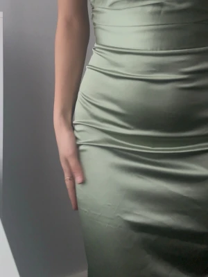 Ljusgrön långklänning i satin - Säljer en elegant ljusgrön långklänning med smala axelband och figurnära passform. Klänningen är tillverkad i ett glansigt satinmaterial som ger en lyxig känsla. Den har sömmar som framhäver midjan och en slät, stilren design utan mönster eller extra detaljer.