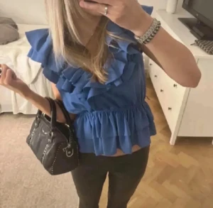 Blå volangblus med omlott - Säljer en blå blus med volanger och omlott-design. Toppen är ärmlös och har en elastisk midja som ger en snygg passform. Materialet känns lätt och luftigt, perfekt för varmare dagar. Volangerna ger en cool och trendig look.