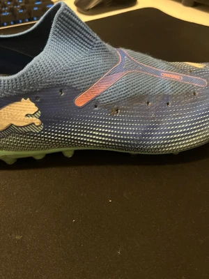 Blå Puma fotbollsskor med strumpdesign - Säljer ett par blå fotbollsskor från Puma med strumpdesign och mönstrad ovandel. Skorna har en ljusgrön sula med fasta dobbar och ett stilrent Puma-logotryck i vitt på sidan. Ovandelen är tillverkad i syntetmaterial och har en tight passform utan snörning.