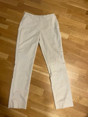 Beiga kostymbyxor från H&M, strl 38 - Snygga beiga kostymbyxor från H&M i storlek 38. De har raka ben, midwaist och klassiska fickor både fram och bak. Materialet är mjukt och känns som bomull blandat med syntet, perfekt för en clean och stilren look.