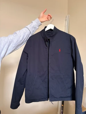 Svart vindjacka från Polo Ralph Lauren - Stilren svart vindjacka från Polo Ralph Lauren med röd broderad logga på bröstet. Jackan har ståkrage, dragkedja och två sidofickor. Materialet är polyester och bomull, perfekt för dig som gillar klassisk och enkel design.
