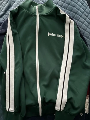 Grön zip hoodie från Palm Angels - Säljer en grön hoodie från Palm Angels med vita och svarta ränder längs ärmarna. Tröjan har hel dragkedja framtill och klassisk logga på bröstet. Materialet är polyester och passformen är loose. Perfekt för streetwear och chill.