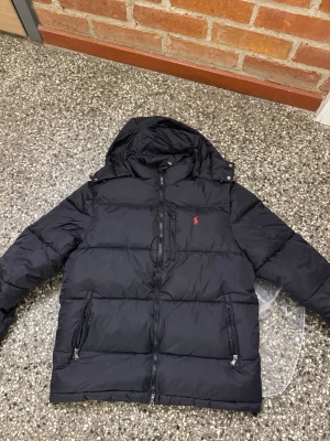 Svart pufferjacka från Polo Ralph Lauren - Svart pufferjacka från Polo Ralph Lauren med huva, dragkedja framtill och två sidofickor med dragkedja. Jackan har det klassiska röda Polo-logot broderat på bröstet och är tillverkad i polyester. Perfekt för kalla dagar.