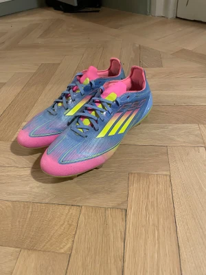 Adidas F50 fotbollsskor - Säljer ett par Adidas F50 fotbollsskor i en riktigt färgstark kombo av blått, rosa och neongult. Skorna har snörning, spetsig tå och platt sula med dobbar för gräsplan och konstgräs. Ovandelen är i syntetmaterial och har de klassiska Adidas-ränderna på sidan. Storlek 41,5✅