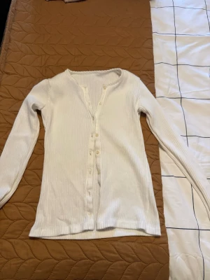 Brandy vit kofta med knappar - Säljer en vit ribbad kofta med lång ärm och knappar framtill. Koftan har en smickrande v-ringad hals och är tillverkad i ett mjukt, stretchigt material som sitter snyggt på kroppen. Perfekt att slänga över en t-shirt eller linne för en chill look.