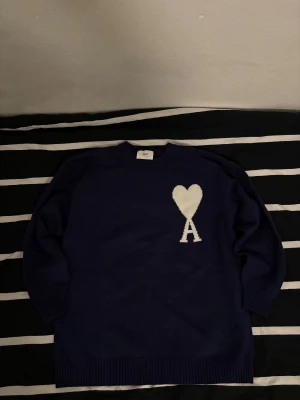 Mörkblå stickad tröja från Ami - Snygg mörkblå stickad tröja från Ami med ett stort vitt hjärta och bokstaven A på bröstet. Tröjan har rund hals, ribbade muddar och lång ärm. Perfekt för dig som gillar stilrena detaljer och vill ha något som sticker ut lite extra. Hör av dig om du har några frågor.