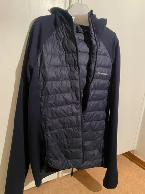 Mörkblå quiltad jacka Peak Performance - Snygg mörkblå jacka från Peak Performance i storlek S. Jackan har quiltad framsida och rygg, huva och dragkedja. Materialet är polyester och syntet, perfekt för kyliga dagar. Stilren design med diskret logga på bröstet.