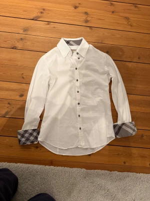 Vit Burberry skjorta - Snygg vit skjorta från Burberry London i storlek S. Skjortan har klassisk krage och knappar framtill. Insidan av kragen och manschetterna har grått och svart rutigt mönster som ger en cool kontrast. Tillverkad i mjuk bomull med lite stretch för extra komfort.