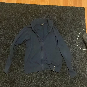 Mörkblå kofta från Indicode storlek S - Säljer en stilren mörkblå kofta från Indicode i storlek S. Koftan har lång ärm, hög krage och hel dragkedja framtill. Materialet är mjukt och känns som bomull, perfekt för lager-på-lager. Diskret logga vid nederkanten och ribbade muddar vid ärmslut och midja.