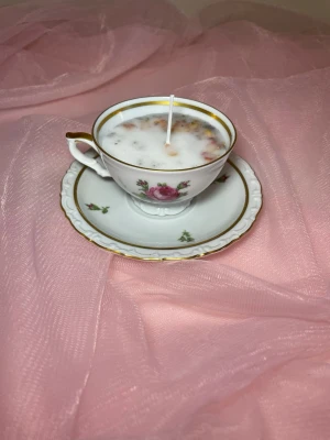 Doftljus i en tekopp i en vit färg med rosa blommor och torkade blommor på nr 2 - Det här ljuset är gjort av återanvändbara material. Tekoppen är köpt secondhand och stearinet är från kyrkor som tänkte slänga det. Det här ljuset har en svag doft utav vanilj, så känsliga även kan använda det. Det finns även fler ljus med denna design på hemsidan, om man vill köpa sets. Det här ljuset har även torkade blommor på. 