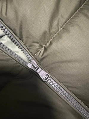 Olivgrön pufferjacka Polo Ralph Lauren - Olivgrön pufferjacka från Polo Ralph Lauren med huva, stora quiltade sektioner och dragkedja framtill. Jackan har en broderad logga på bröstet och flera praktiska fickor med dragkedja. Materialet är polyester och jackan är perfekt för kalla dagar.