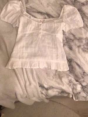 Vit blus med volang från Nelly - Supersöt vit blus från Nelly i storlek 36. Blusen har korta puffärmar, knappar framtill och volangkant längst ner. Tunt randigt mönster i tyget och smock vid halsen ger en romantisk vibe. Perfekt för vår och sommar!