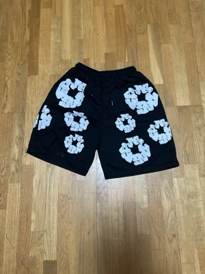 Svarta shorts African Diaspora Denims - Svarta shorts från African Diaspora Denims med vita blommiga mönster över hela plagget. De har elastisk midja med snörning och en avslappnad passform. Perfekta för sommaren och streetwear-stil.
