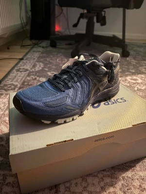 Asics Skor - Säljer ett par snygga oanvända blå Asics sneakers med svart och vit detalj. Skorna har mesh- och syntetmaterial, rund tå och platt sula. De har snörning med mönstrade skosnören och en sportig look, perfekt för dig som gillar stil och komfort.