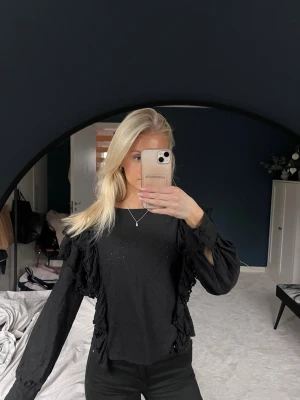 Svart blus med volangdetaljer - Säljer en svart blus med långa ärmar och volangdetaljer längs axlar och sidor. Perfekt för dig som gillar en stilren men ändå unik look.