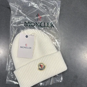 Vit ribbstickad mössa från Moncler - Vit ribbstickad mössa från Moncler med klassisk uppvikt kant och broderad logotyp framtill. Tillverkad i mjukt material som håller dig varm och snygg under kyliga dagar. Perfekt accessoar för en clean och stilren look.