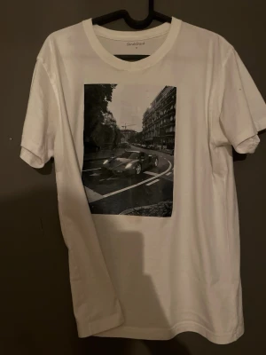 Vit t-shirt med biltryck från GardeRobe - Vit t-shirt från GardeRobe med ett stort svartvitt tryck av en sportbil och stadsmiljö på bröstet. Klassisk rund halsringning och korta ärmar. Perfekt för dig som gillar streetstyle och snygga prints.