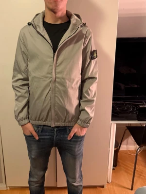 Beige vindjacka från Stone Island - Säljer en beige vindjacka från Stone Island med huva och dragkedja framtill. Jackan har svarta detaljer, elastiska muddar och klassisk Stone Island-patch på vänster ärm. Perfekt för dig som vill ha en stilren och sportig look.