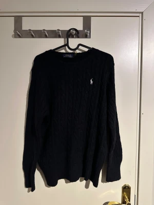 Svart kabelstickad tröja Polo Ralph Lauren - Svart kabelstickad tröja från Polo Ralph Lauren i storlek L. Klassisk design med rund hals och vit broderad logga på bröstet. Tröjan har långa ärmar och är gjord i mjukt material som passar perfekt för kyliga dagar.