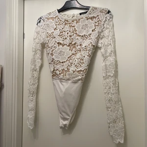Vit spetsbody från Boohoo - Säljer en vit body från Boohoo med långärmade spetsdetaljer och stora blommor i spetsen. Toppen har öppen rygg med knyt och rund halsringning. Nederdelen är slät och har praktisk knäppning. Perfekt för dig som vill ha något extra till din outfit.