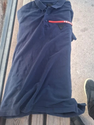 Marinblå polotopp från Prada - Säljer en marinblå polotopp från Prada med klassisk krage och knappar framtill. Toppen har en röd detalj med Prada-logga på bröstet och är tillverkad i mjuk bomull. Perfekt för dig som gillar stilrena designer och exklusiva märken.