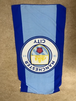 Manchester City badhandduk blå - Snygg badhandduk med Manchester Citys klubbmärke i mitten. Handduken har breda blå och ljusblå ränder och är tillverkad i mjuk frotté. Perfekt för stranden eller poolen för dig som älskar fotboll och vill visa ditt lag.