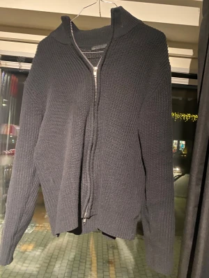 Svart stickad kofta med dragkedja - Säljer en svart stickad kofta med grov ribbad struktur och hög krage. Koftan har en hel dragkedja framtill och långa ärmar, perfekt för kyliga dagar. Enkel och stilren design som funkar till många olika outfits. Leverera snabbt