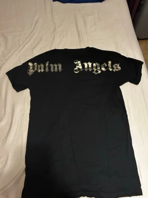 Svart Palm Angels x Moncler t-shirt - Svart t-shirt från Palm Angels x Moncler med guldiga tryck på både fram- och baksidan. Framsidan har Moncler-logga och ett stort 'P' i guld, medan baksidan har Palm Angels-tryck i metallic. Klassisk rund hals och korta ärmar. Perfekt för dig som gillar streetwear och exklusiva samarbeten.