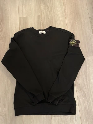Svart Stone Island sweatshirt - Svart långärmad sweatshirt från Stone Island med klassisk rund hals och ikonisk logotyp-patch på vänster ärm. Tröjan är i mjuk bomull och har ribbade muddar vid ärmslut och nederkant. Perfekt för dig som gillar streetwear och snygga detaljer.