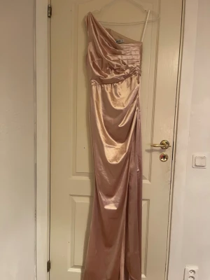 Rosa långklänning - Säljer en elegant långklänning i rosafärgad satin. Klänningen har oneshoulder-design med draperad överdel och slits i sidan. Materialet är glansigt och mjukt, perfekt för dig som vill sticka ut med en lyxig look. Dragkedja i ryggen för enkel påklädning. Har en hög slit på framsidan.