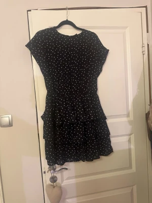 Svart prickig volangklänning - Säljer en svart kortklänning med vita prickar och volangdetaljer. Klänningen har rund hals, är kortärmad och har flera lager med volanger som ger en snygg och lekfull look. Perfekt för dig som gillar mönstrade plagg med lite extra flärd.